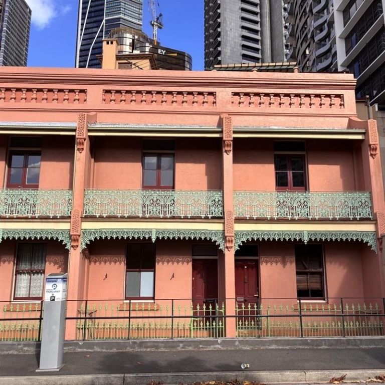 A two-story pink building with green ironwork balconies, set against a backdrop of modern high-rise buildings on a cloudy day.