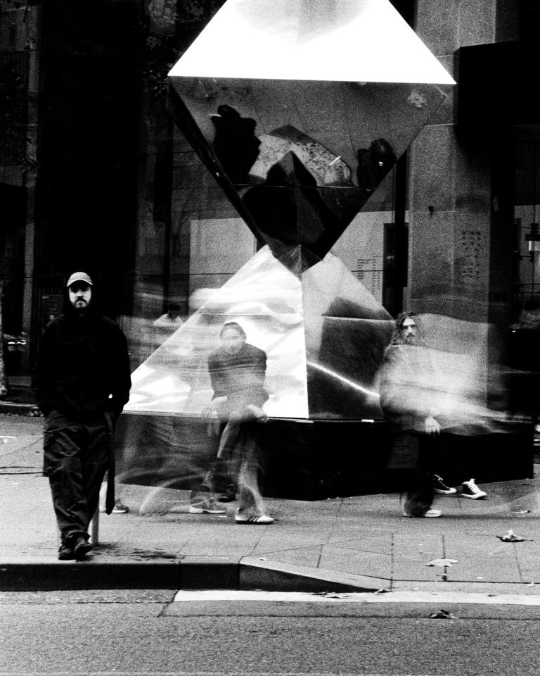 Black-and-white long-exposure street photo of blurred pedestrians around a reflective geometric sculpture, with one man standing at left.