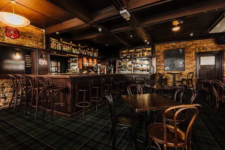 Cozy pub interior with wooden furniture, a well-stocked bar, brick walls, and warm lighting, creating an inviting atmosphere.
