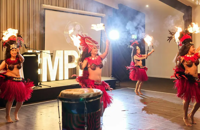 Performers in traditional Polynesian attire dance with flaming torches on a stage, with large illuminated letters and a drum in the background.
