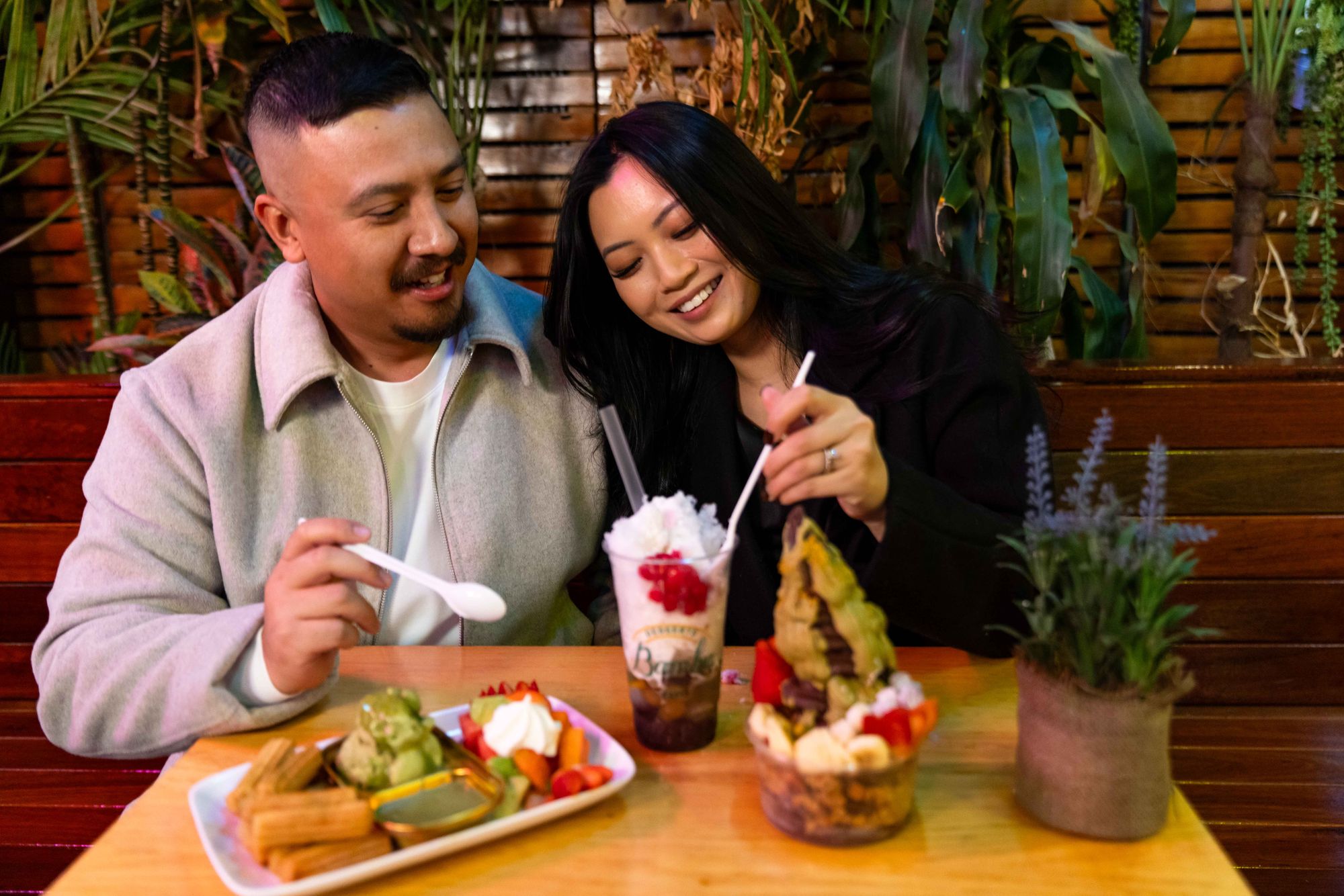 A smiling couple enjoys a dessert table with ice cream, shaved ice, and fruit, surrounded by plants in a cozy setting.