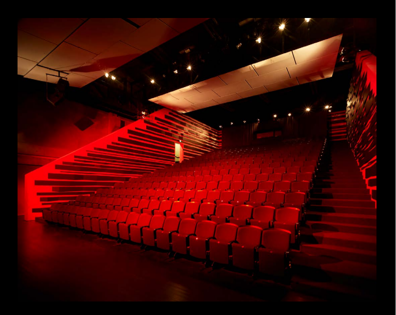 A wide-angle interior photograph of an empty theatre auditorium featuring steeply tiered seating. The room is bathed in dramatic red lighting, which intensely highlights the rows of red velvet seats and the angular architecture of the side walls and staircases. Above, white acoustic panels hang from the ceiling, reflecting the light.