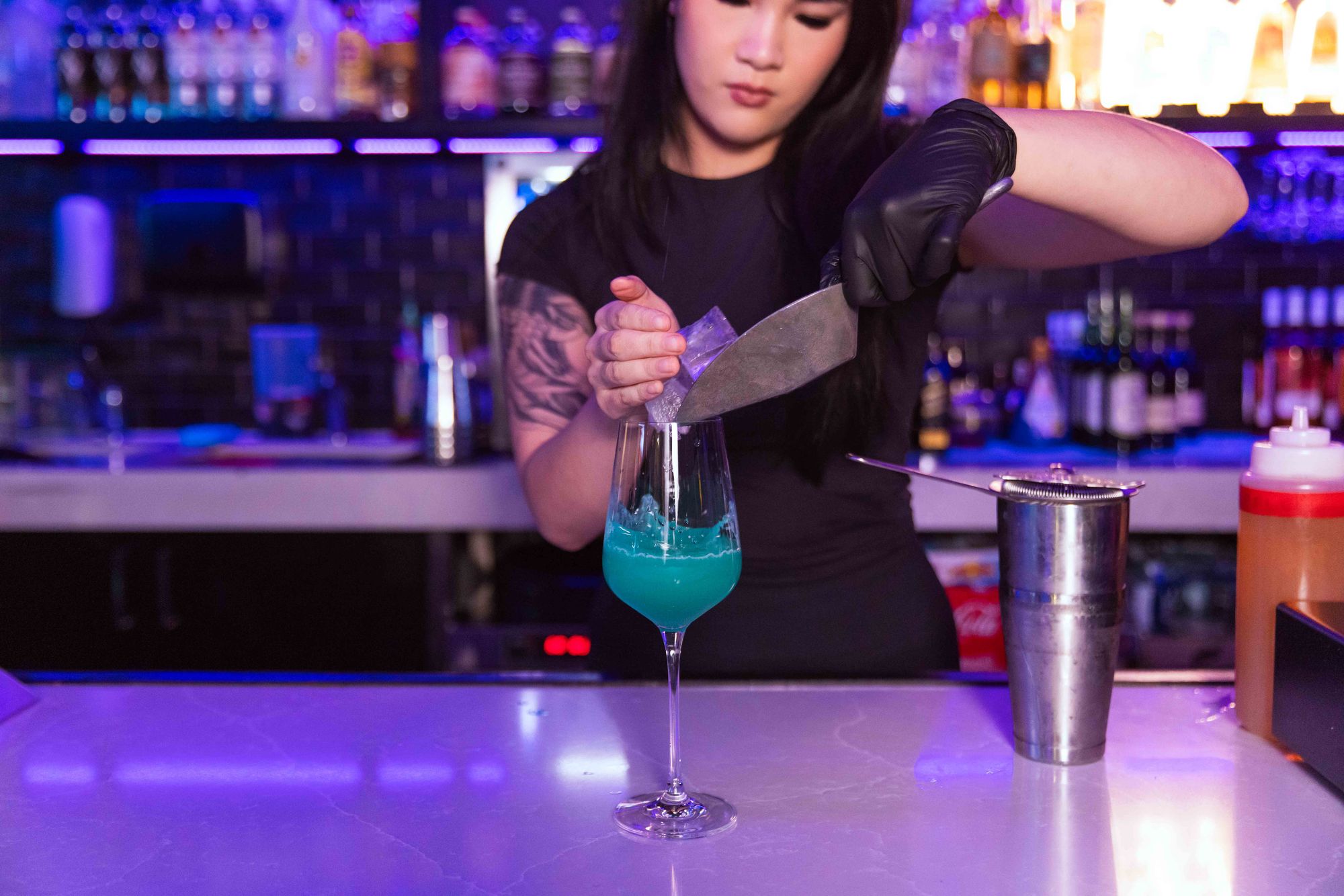 Bartender with tattoos and gloves prepares a blue cocktail at a bar, using a large knife to add ice. Shelves with bottles in the background.