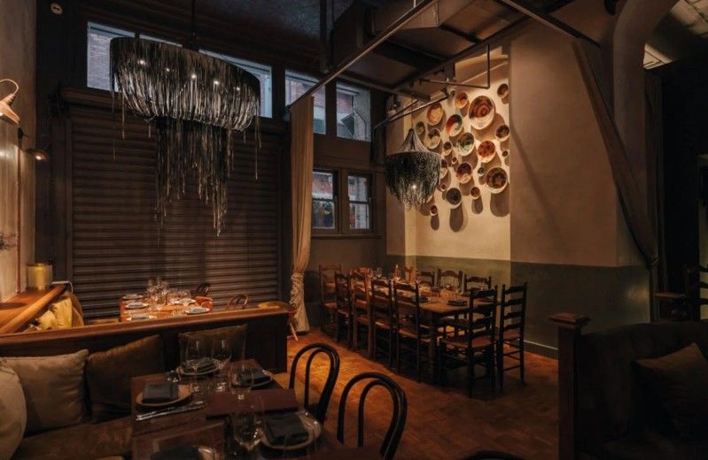 Cozy restaurant interior with warm lighting, wooden tables, and a wall art installation featuring various plates. Large decorative chandeliers hang above.
