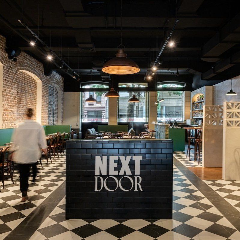 Modern restaurant interior with checkered floor, exposed brick wall, and a counter with "Next Door" sign. Warm lighting and wooden tables enhance ambiance.