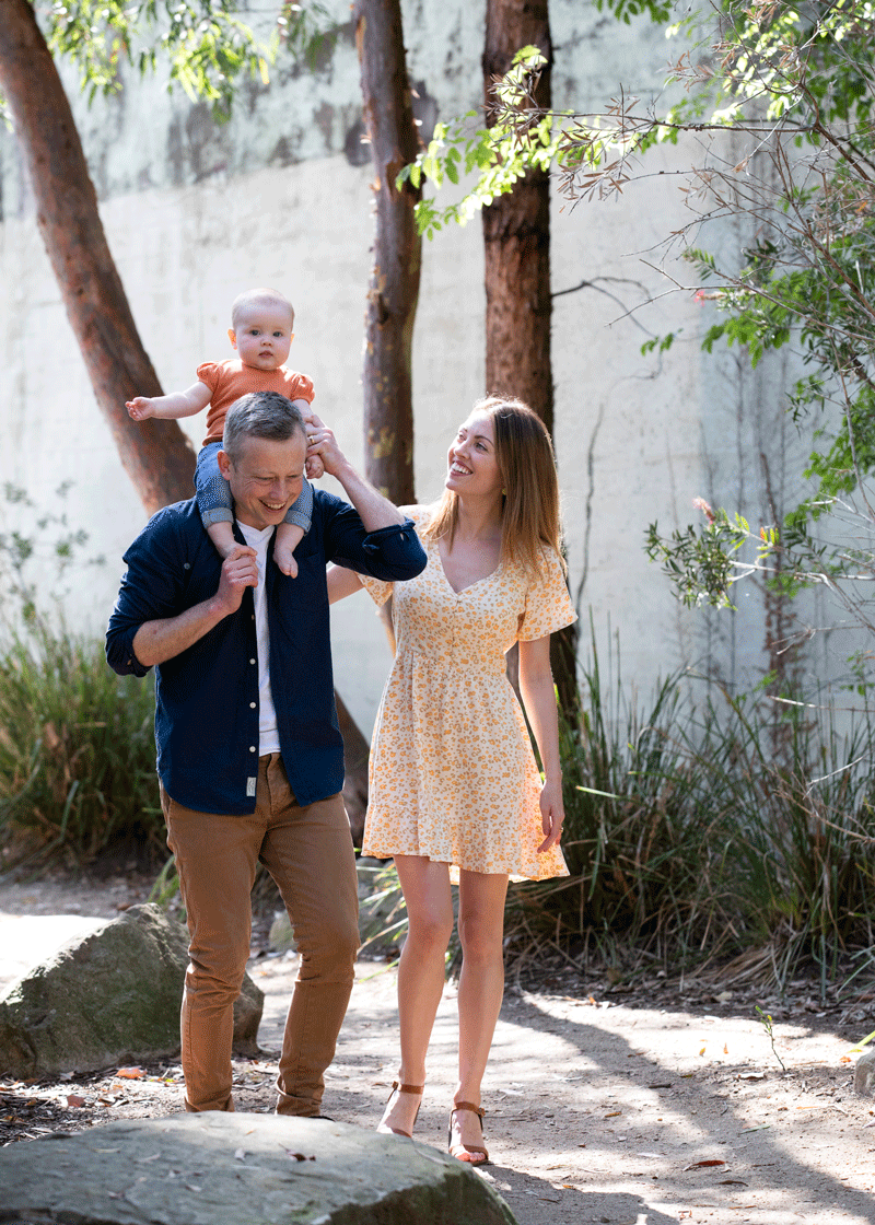 A family are walking together in the park. The father is carrying their child on his shoulders
