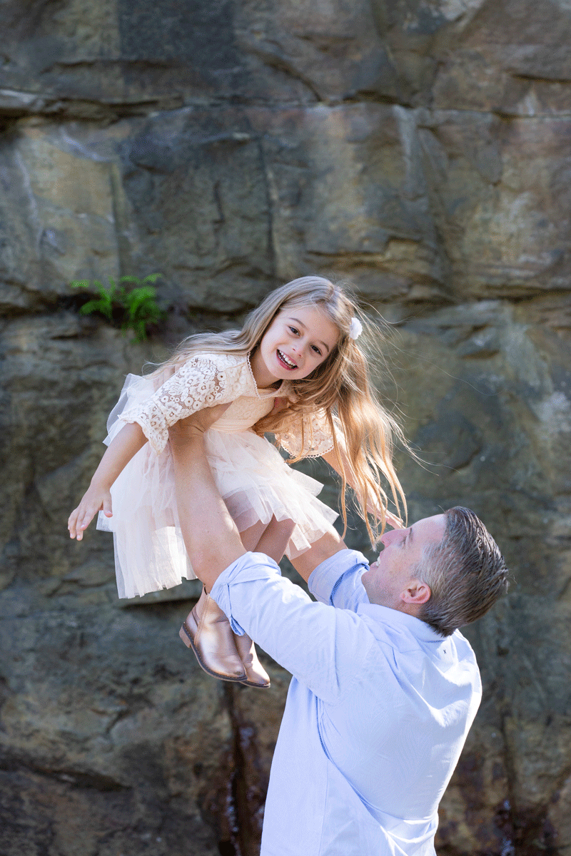 A father and his daughter are having their photos taken. The father is holding his daughter up in the air above him and she is smiling