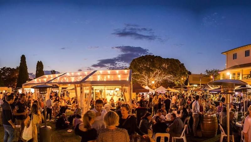 Outdoor evening festival with a lively crowd under string lights, surrounded by trees and tents against a dusk sky.