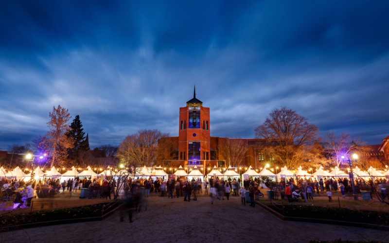 Festive outdoor market at dusk with bright lights, a crowd of people, and a tall, illuminated brick building under a deep blue sky.