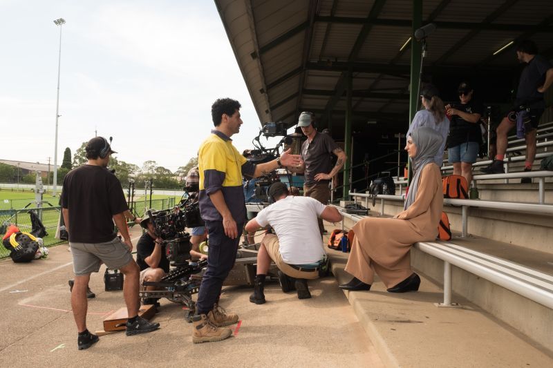 Film crew recording a scene at outdoor bleachers, with a camera setup. A woman in a hijab and beige dress sits while people collaborate nearby.