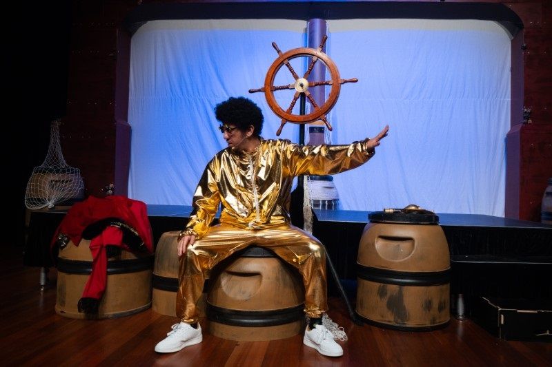 Person in a shiny gold outfit poses dramatically on a stage, sitting on barrels near a ship wheel, with dramatic lighting.