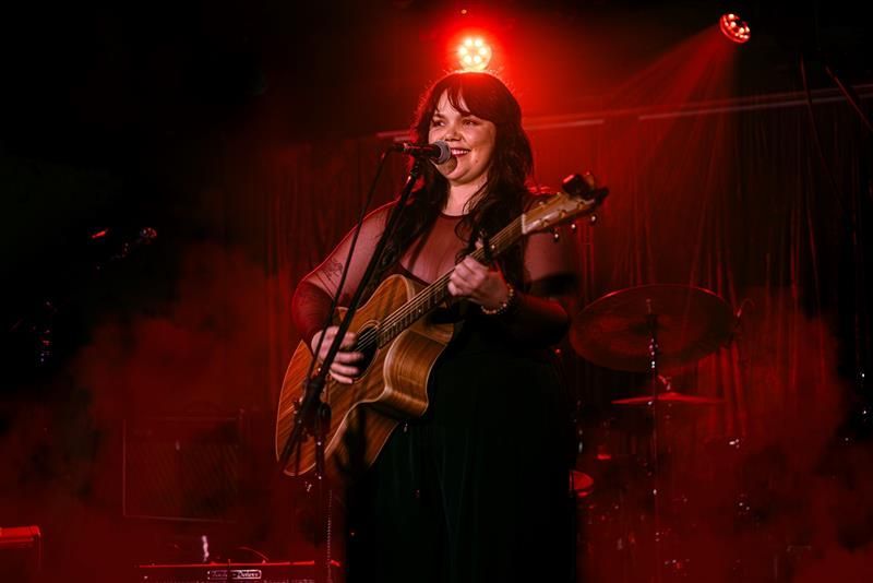 A woman plays an acoustic guitar and sings on stage, surrounded by red lighting and smoke, creating an atmospheric performance setting.