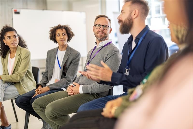 Diverse group of people in a discussion, seated in a circle in a brightly lit room, with a whiteboard in the background.