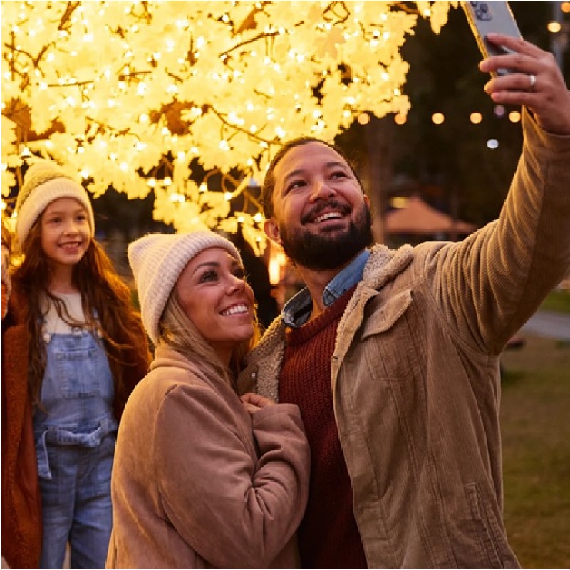 A family taking a selfie under a brightly lit tree adorned with glowing lights.