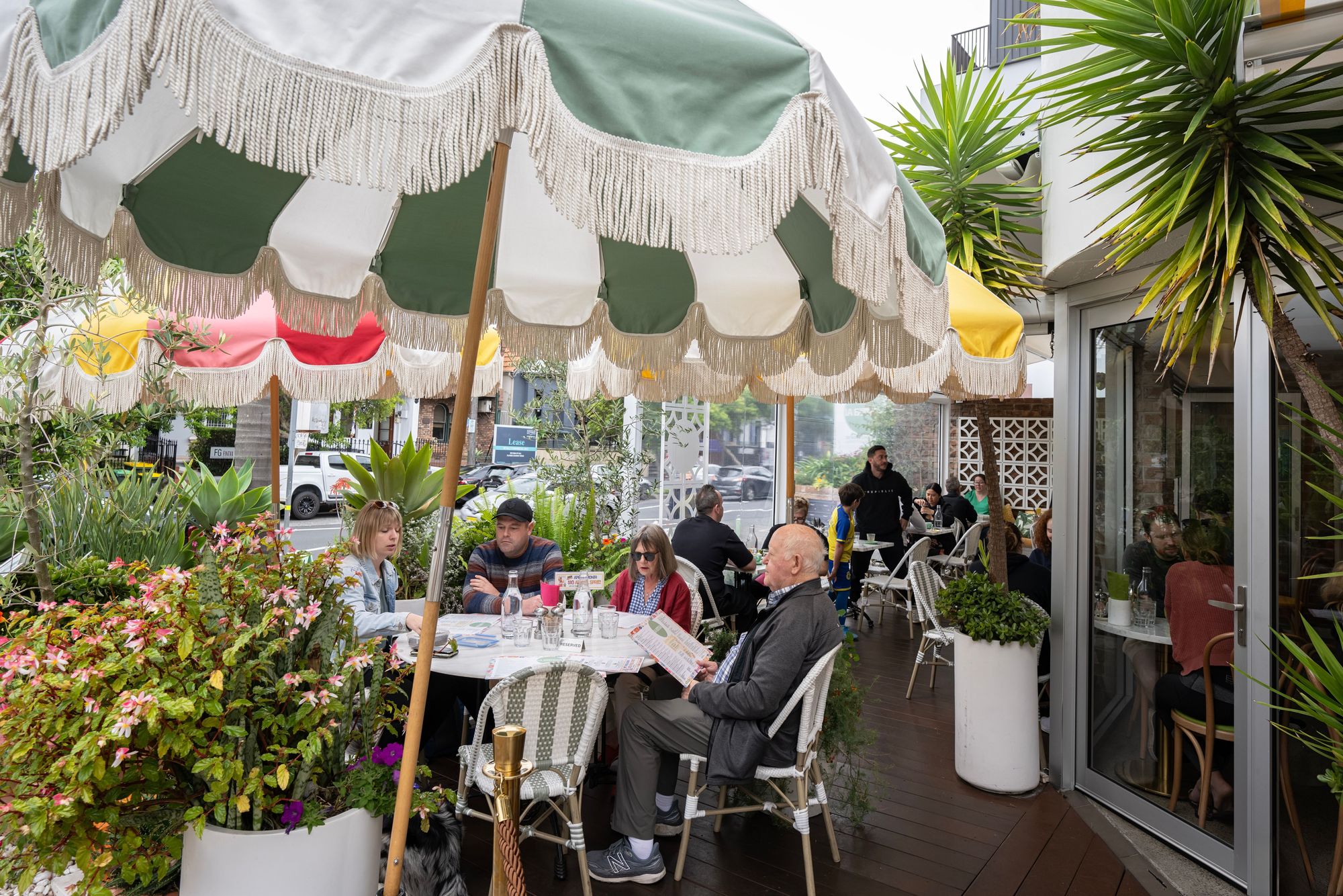 People dining at an outdoor café with colourful umbrellas, surrounded by greenery, on a wooden deck.