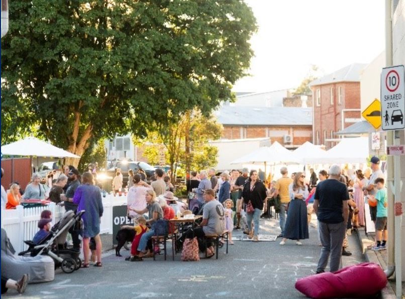 A lively outdoor market scene with people socialising, sitting at tables, and walking. Stalls and trees are visible under a clear sky.