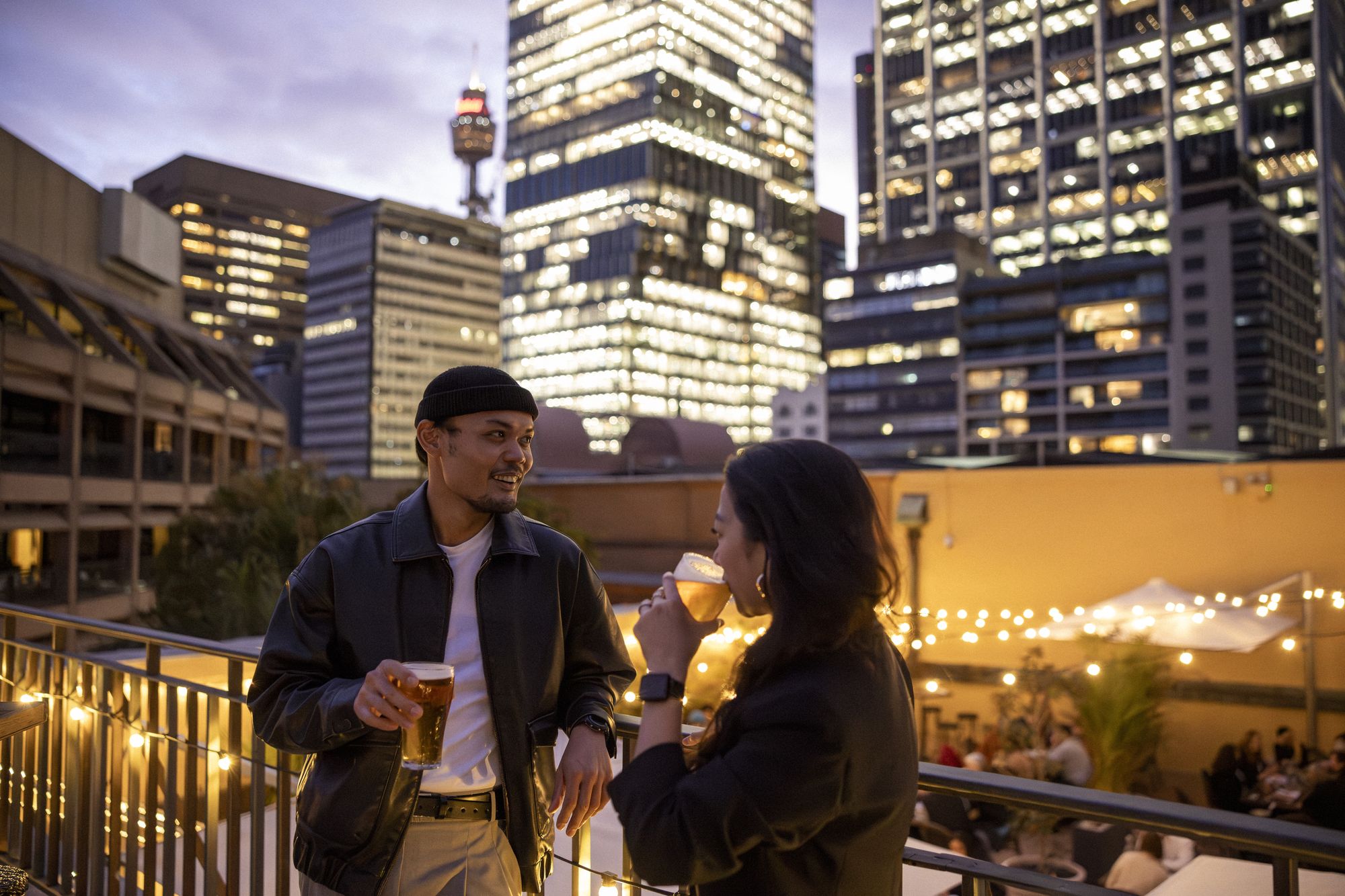 Two people enjoying drinks on a rooftop at dusk, with city skyscrapers and string lights in the background.