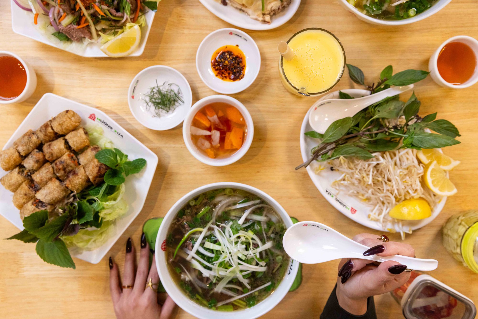 A table with Vietnamese dishes including pho, spring rolls, fresh herbs, dipping sauces, and a mango smoothie. Hands holding a spoon and chopsticks.