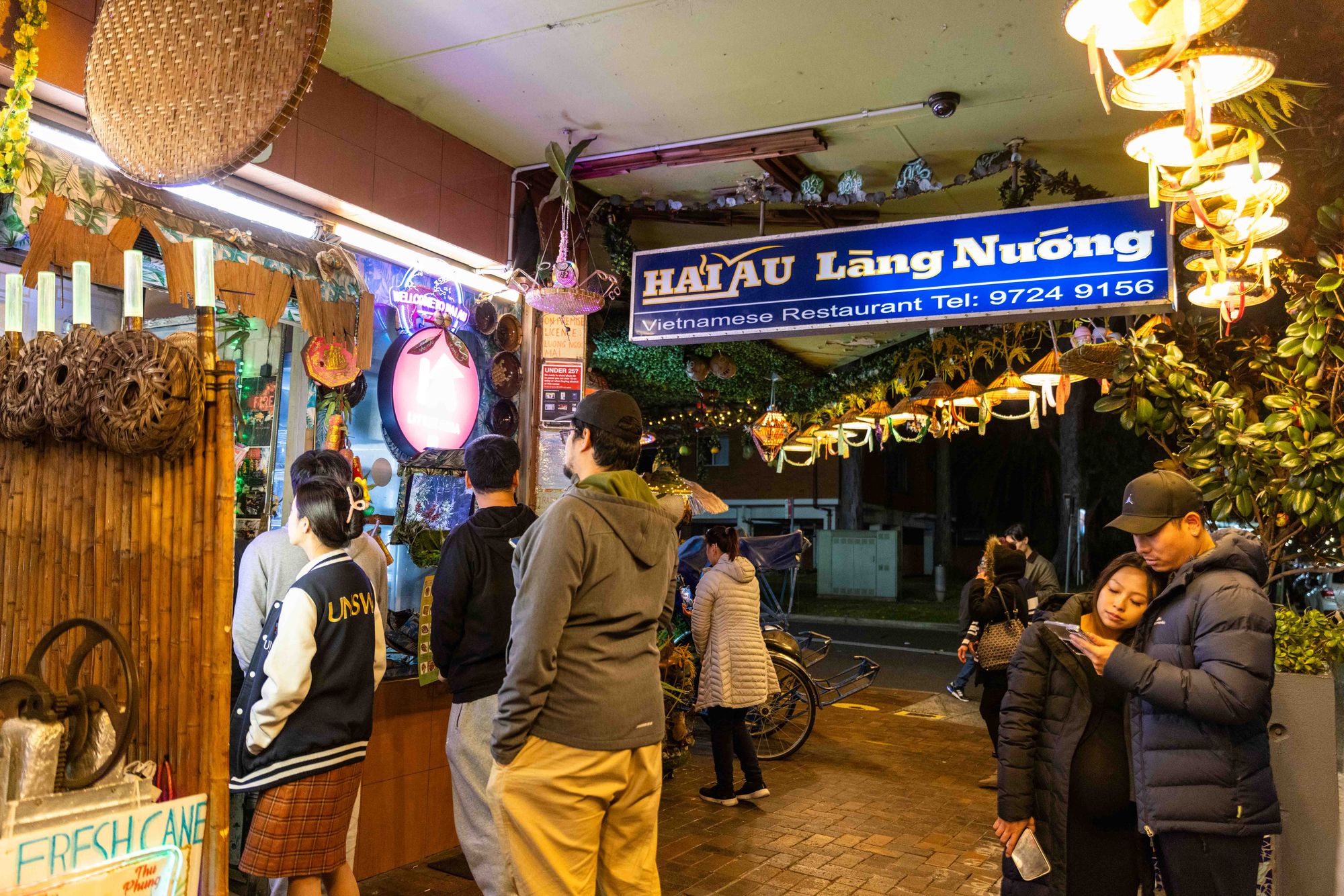 People waiting outside a Vietnamese restaurant at night, with a sign reading "Hai Au Làng Nướng" and a display of hanging decorations.