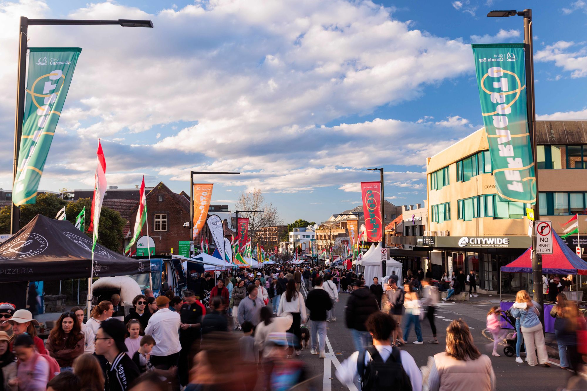 A bustling street festival with colorful banners, tents, and a crowd of people under a partly cloudy sky.