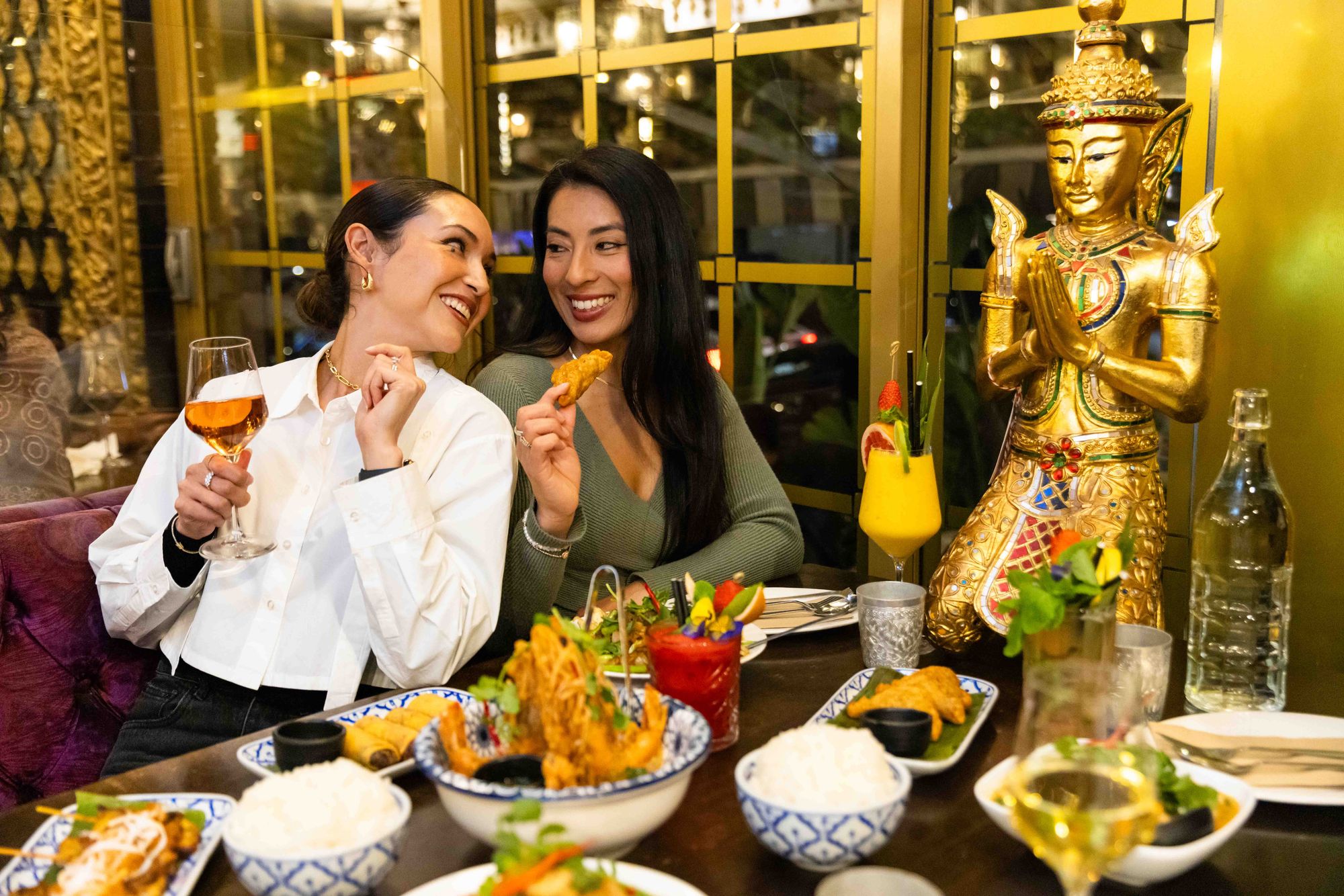 Two women enjoying drinks and appetizers at a restaurant, seated beside a golden statue, with various dishes and drinks on the table.