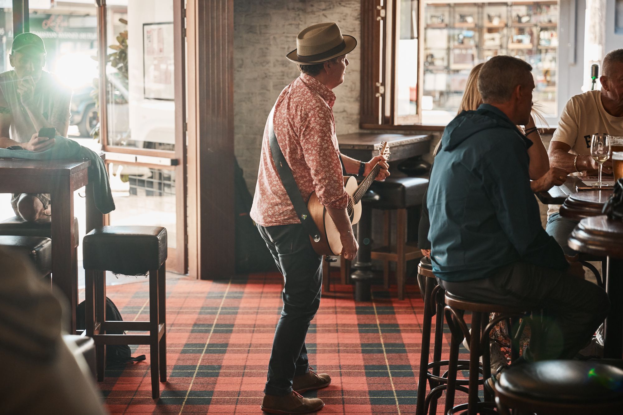 A musician in a hat and patterned shirt plays guitar in a cozy bar with patrons seated around. Sunlight streams through the entrance.
