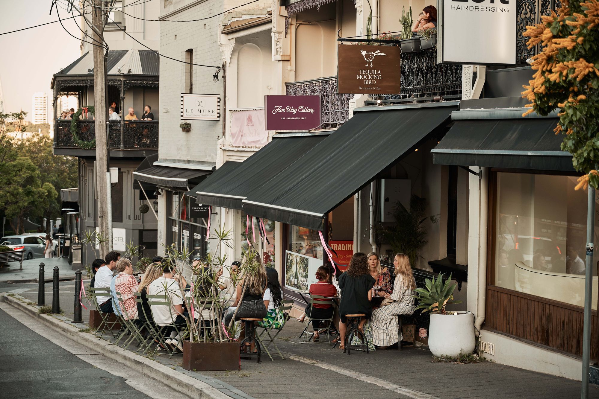 People sitting at outdoor tables of a café on a quiet street, with some greenery and buildings in the background on a sunny day.