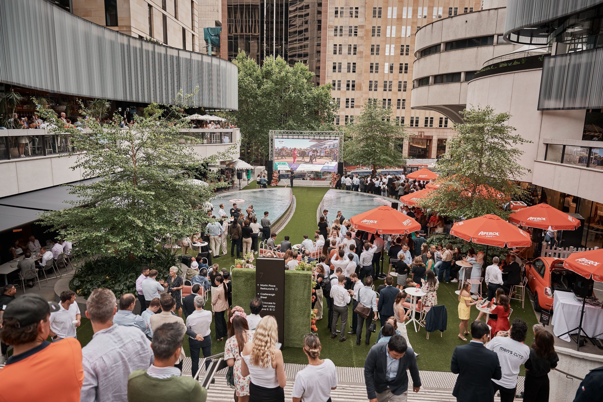 A lively outdoor event with people gathered around a large screen. Bright orange umbrellas and surrounding modern buildings enhance the scene.