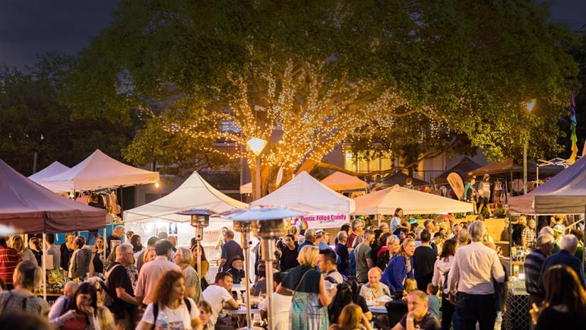 A bustling night market with people mingling under illuminated trees and white canopies, creating a lively and festive atmosphere.