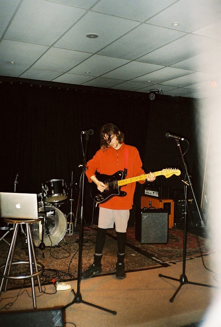 The artist is on stage, dressed in a red quarter zip with checkered shorts layered over black running tights with black socks and black Nike running shoes. They are playing guitar standing and looking down in front of some band music equipment accompanied by a laptop on a stool playing the backing track. It is quite bright due to the flash of the camera. Photograph taken on a real film camera.