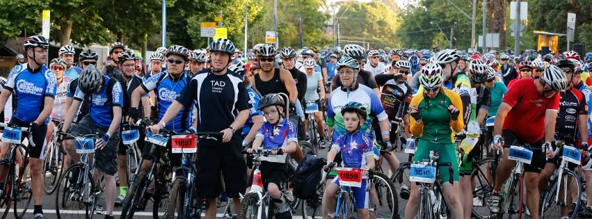 A large group of cyclists at the starting line of an event, wearing helmets and various colorful jerseys, with trees in the background.