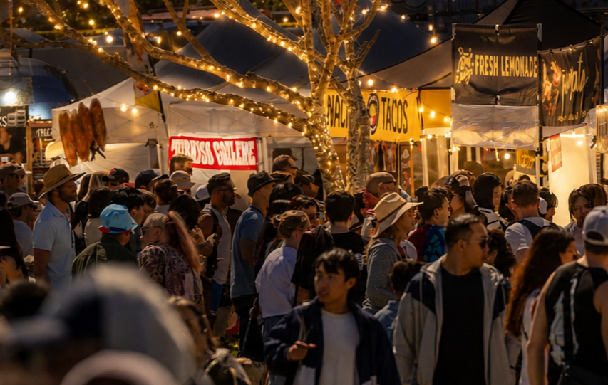 Crowded outdoor night market with people browsing food stalls under string lights, featuring churros, tacos, and fresh lemonade signs.