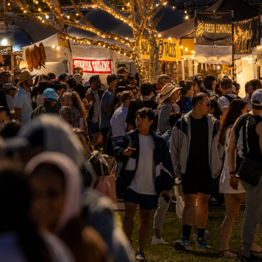 A lively night market with people browsing food stalls under string lights, featuring tacos and lemonade stands.