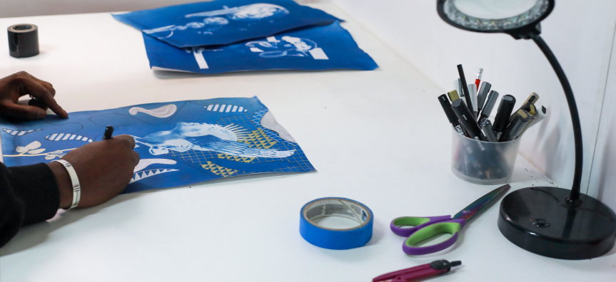 A person drawing on blue paper at a desk with scattered art supplies, including scissors, tape, pens, and a desk lamp.
