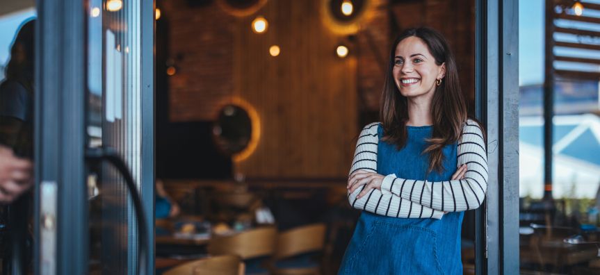 Smiling woman in a striped shirt and blue dress stands with arms crossed in a cozy cafe, with warm lighting and wooden accents in the background.