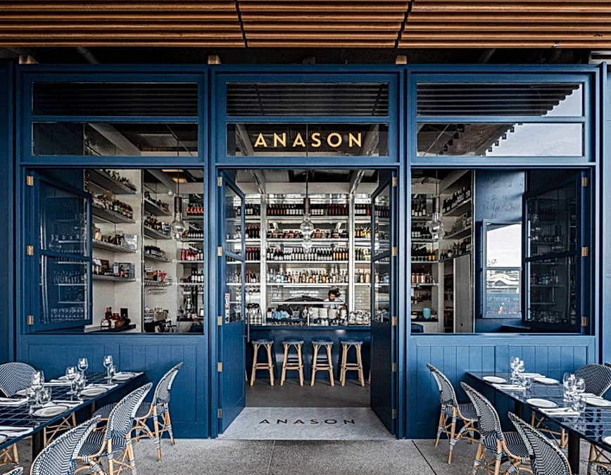 Blue-framed restaurant entrance with "Anason" sign, open doors revealing a stocked bar and wooden stools, surrounded by outdoor dining tables and chairs.