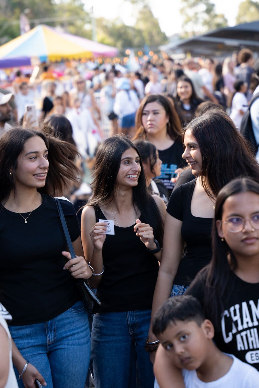 A group of people smiling and walking at an outdoor event, with colorful tents and a bustling crowd in the background.