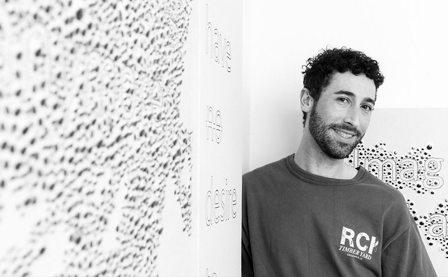 Man with curly hair smiling, leaning against a wall with abstract text art. Wearing a sweatshirt with "RCV" on it. Black and white image.