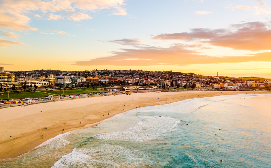 Aerial view of a Bondi Beach at sunset, with gentle waves, scattered people, and a town in the background under a colorful sky.