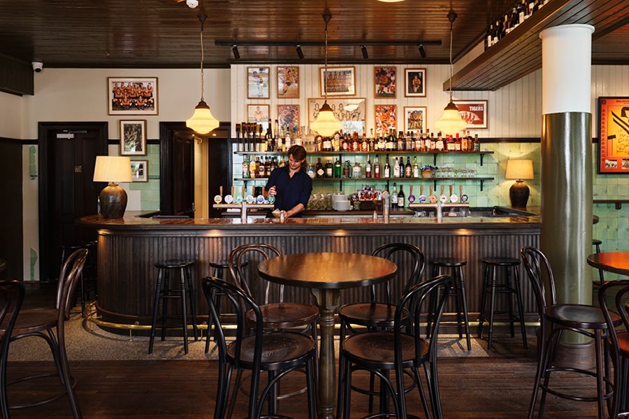 A cozy bar with a wooden counter, stools, pendant lights, and various bottles on shelves. A person stands behind the bar.