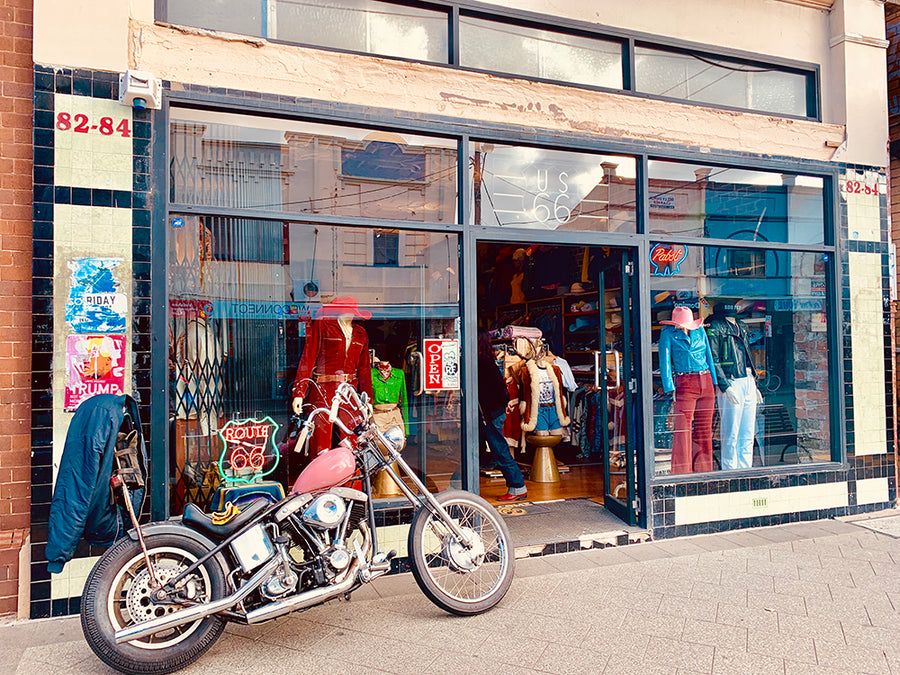 Vintage store with glass front, showcasing mannequins and clothing. A classic motorcycle is parked outside on the sidewalk.