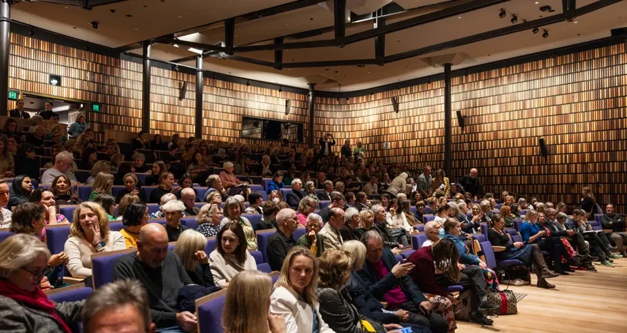 A packed auditorium with people seated, featuring a wood-paneled wall and a high ceiling. Attendees appear engaged and waiting for an event.