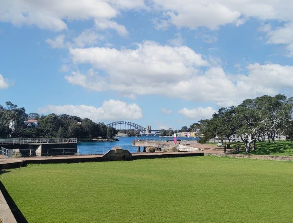 A lush green lawn extends towards a harbor, with a distant bridge under a partly cloudy blue sky. Trees line the right side of the scene.