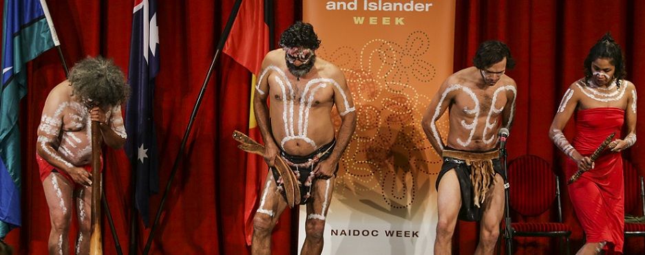 Four Indigenous Australian performers in traditional attire and body paint stand on stage during NAIDOC Week, with flags and a red curtain behind.