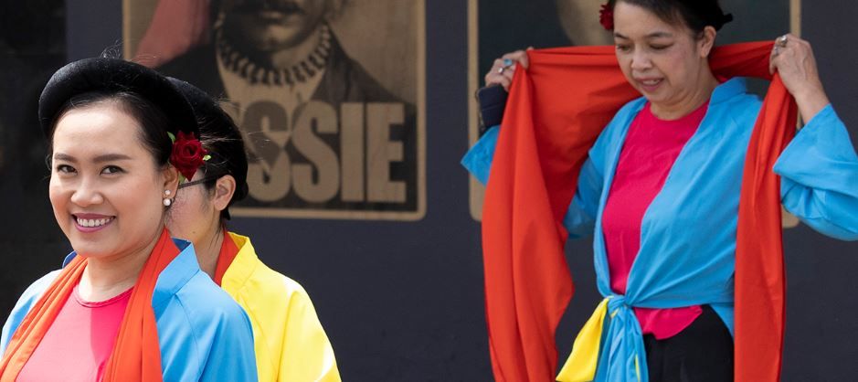 Women in colorful traditional clothing, with blue and yellow attire, red scarves, and floral headpieces, smiling and preparing for a performance.
