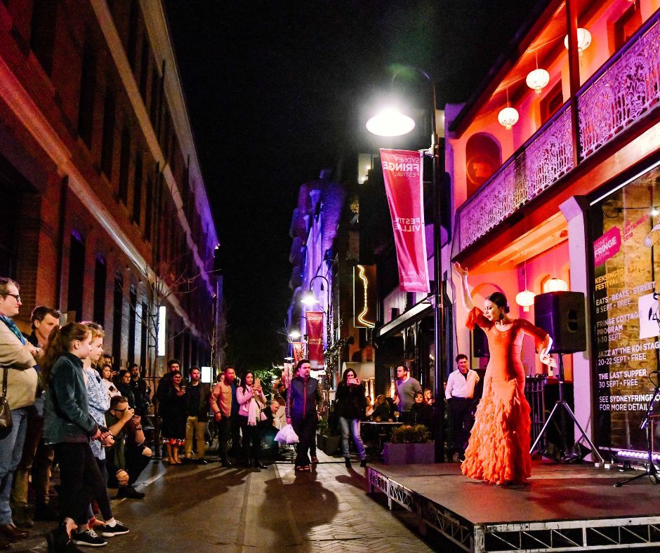 Street performance at night featuring a dancer in a vibrant red dress on a small stage, with an engaged crowd and illuminated buildings in the background.
