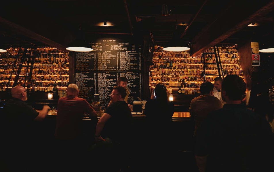 Dimly lit bar with people sitting at the counter, illuminated shelves of bottles in the background, and a large chalkboard menu on the wall.