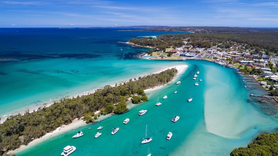 Aerial view of coastal town Huskisson with turquoise waters, white sandy beaches, and boats anchored near shore under a clear blue sky.