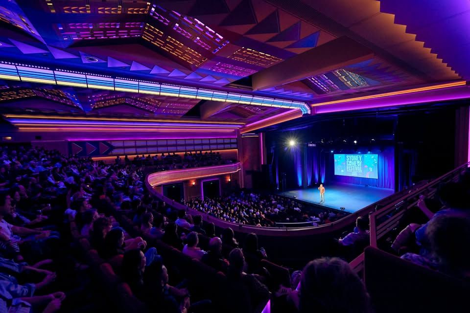 A vibrant theatre with a packed audience, illuminated in purple and blue lights, watches a speaker on stage during a presentation or show.