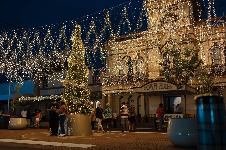 Town hall adorned with festive lights at night, with a Christmas tree and people strolling beneath twinkling decorations.